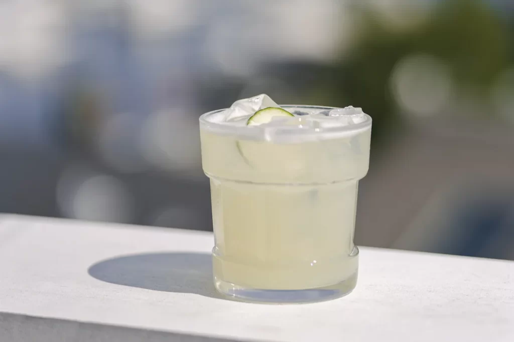 A pale lime cocktail over ice with a slice of lime, on a sunlit white ledge