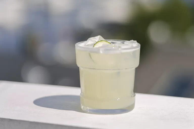 A pale lime cocktail over ice with a slice of lime, on a sunlit white ledge