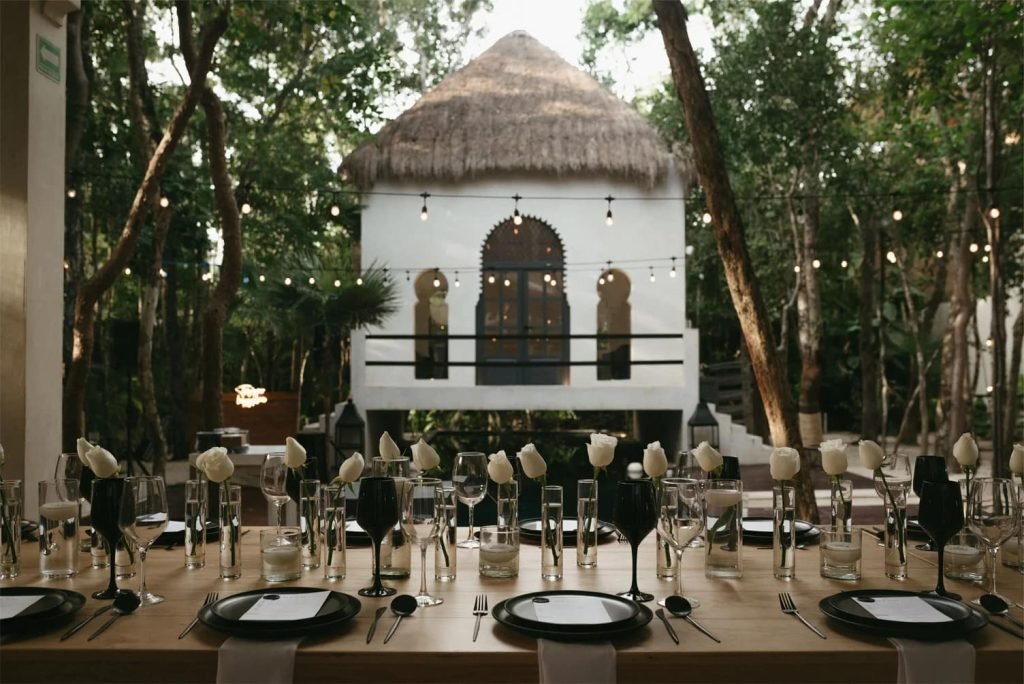 Long table set for a private dinner with stemware and white roses in front of a palapa-roofed villa in the Tulum jungle