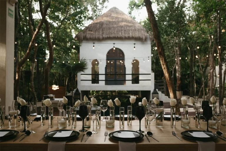 Long table set for a private dinner with stemware and white roses in front of a palapa-roofed villa in the Tulum jungle