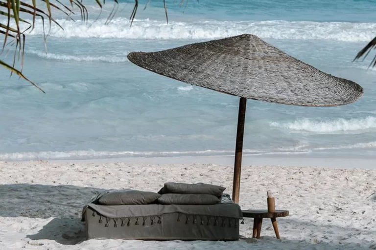 A cushioned day bed under a palm-thatch umbrella on the sand at a Tulum beach club, with the sea behind it