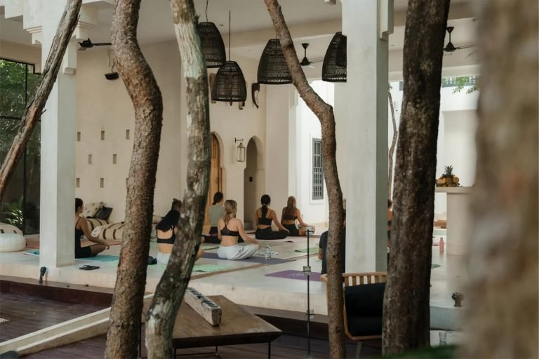 Yoga class seated on a white terrace with arched doorways and woven pendant lamps, seen between tree trunks