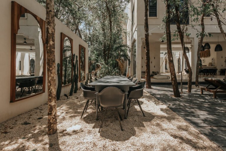 Outdoor dining table on gravel under trees, beside a wall of arched wooden-framed mirrors and a deck with sun loungers