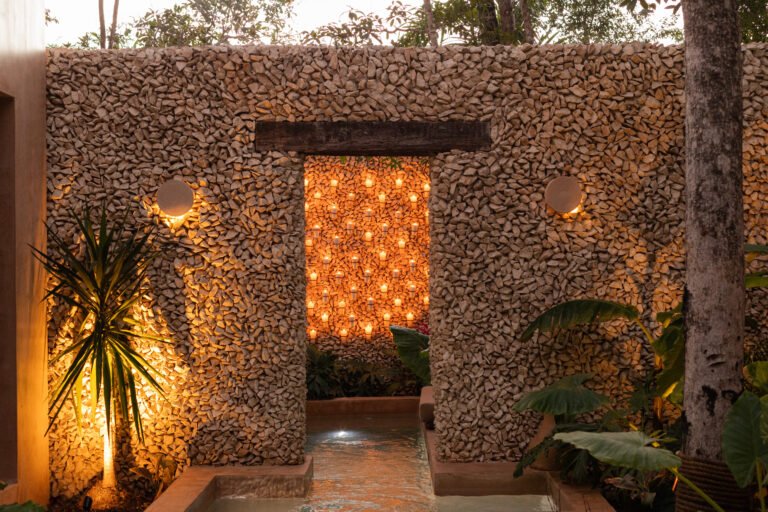 Stone wall lit at dusk, its doorway framing rows of warm lights above a lit water channel and tropical plants