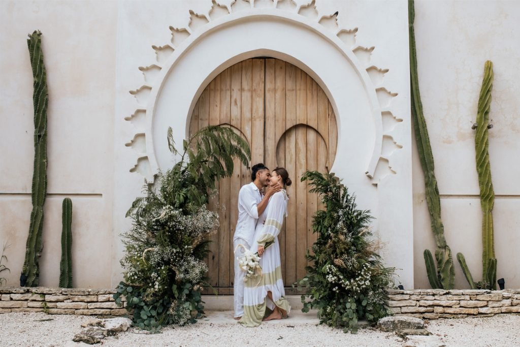 Couple embracing in front of a carved Moorish archway framed by tropical greenery and cacti at a wedding venue in Tulum