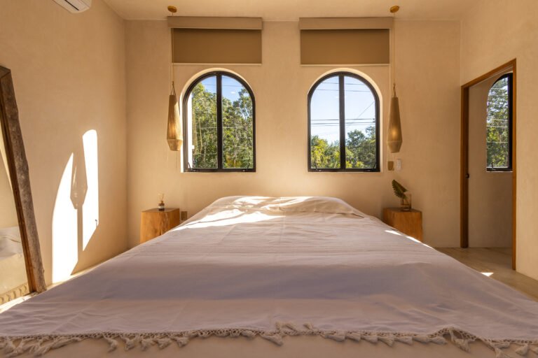 Bedroom at Casa Alba villa with a white bed, woven pendant lamps and two arched windows looking onto treetops